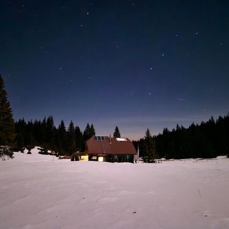 Ein Hütte mit Solarmodulen auf dem Dach, bei Nacht beleuchtet, ist von Schnee und Kiefern umgeben unter einem sternenklaren Himmel.