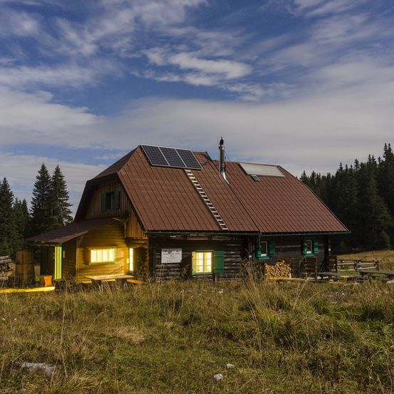 Eine Holzhütte mit braunem Dach und Solarpanelen steht auf einer Wiese unter einem bewölkten Himmel. Bänke und ein Holzstapel sind im Vordergrund.