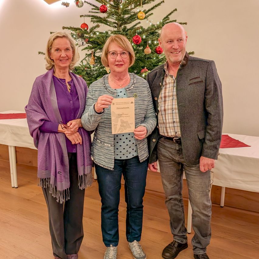 Drei Personen stehen eng beieinander vor einem Weihnachtsbaum und lächeln für ein Foto. Die Frau in der Mitte hält ein Zertifikat. Dahinter steht ein Tisch mit weißen Tischdecken und einem festlich geschmückten Weihnachtsbaum.