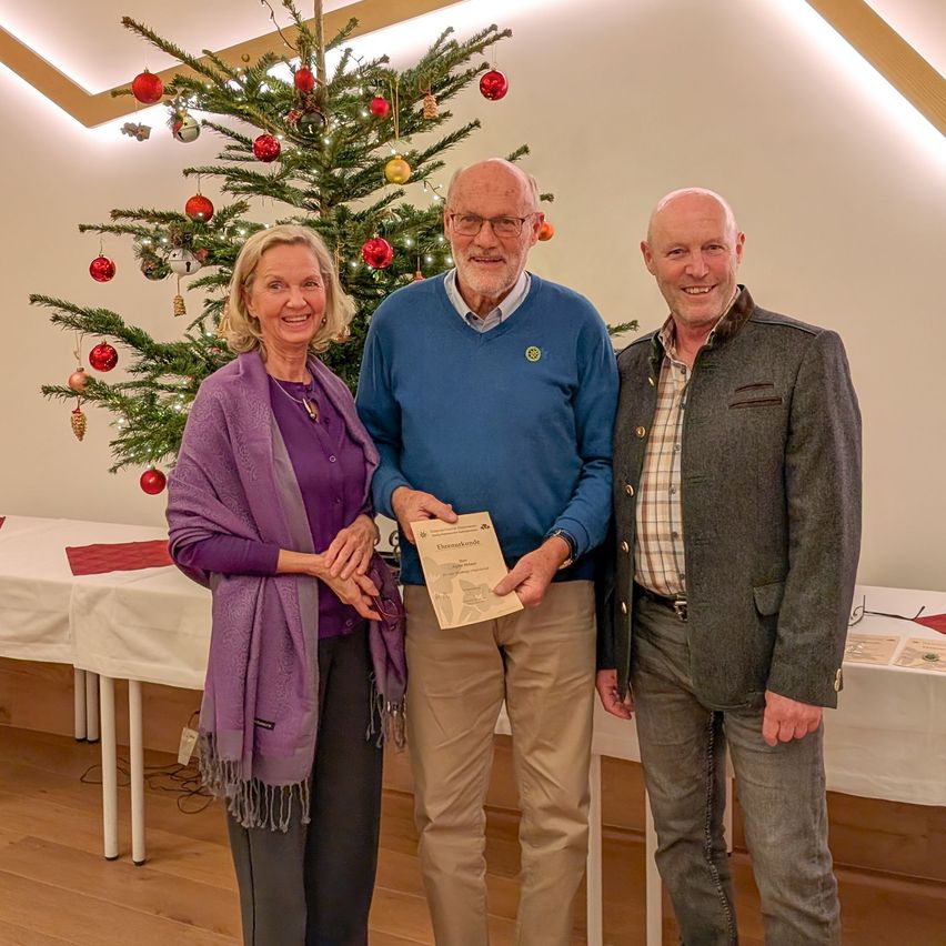 Drei Menschen stehen zusammen vor einem geschmückten Weihnachtsbaum und lächeln für ein Foto. Ein Mann hält eine Urkunde, während eine Frau in einem Schal und ein Mann in einem Mantel ihn flankieren. Dahinter steht ein Tisch mit einer weißen Tischdecke und einer roten Serviette.