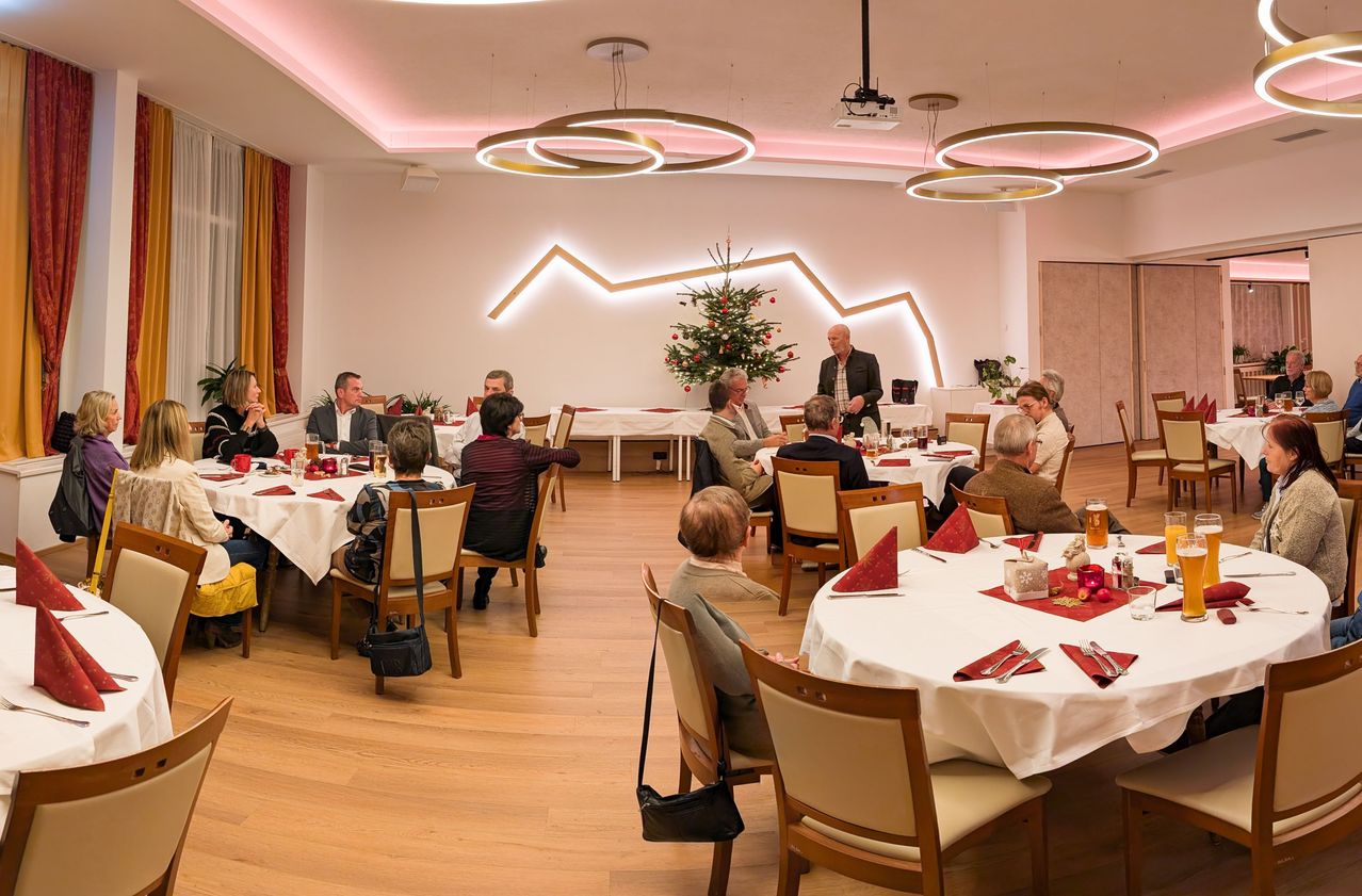 Ein Speisesaal mit Tischen und Stühlen für eine Mahlzeit vorbereitet. Ein Mann steht an einem Tisch mit einem Weihnachtsbaum, und andere sitzen an Tischen. Der Raum hat Holzböden und kreisförmige Lichter.
