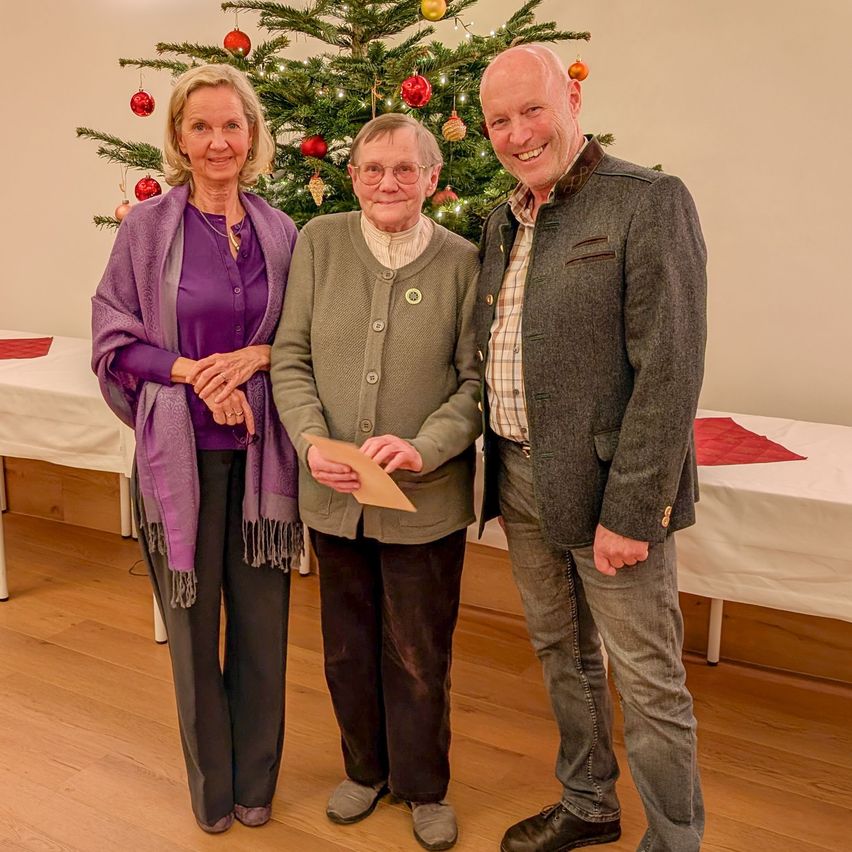 Drei Menschen stehen nebeneinander vor einem geschmückten Weihnachtsbaum und lächeln für die Kamera. Die Frau links trägt einen lila Schal. Die Frau in der Mitte hält ein Papier und der Mann rechts trägt einen grauen Mantel. Hinter ihnen befinden sich Tische, die mit weißen Tüchern und roten Servietten bedeckt sind.