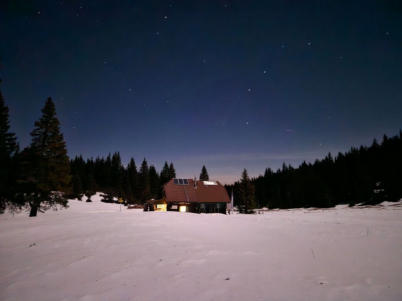 Eine gemütliche Hütte in den verschneiten Bergen bei Nacht mit einem Sternenhimmel, umgeben von hohen Kiefern.