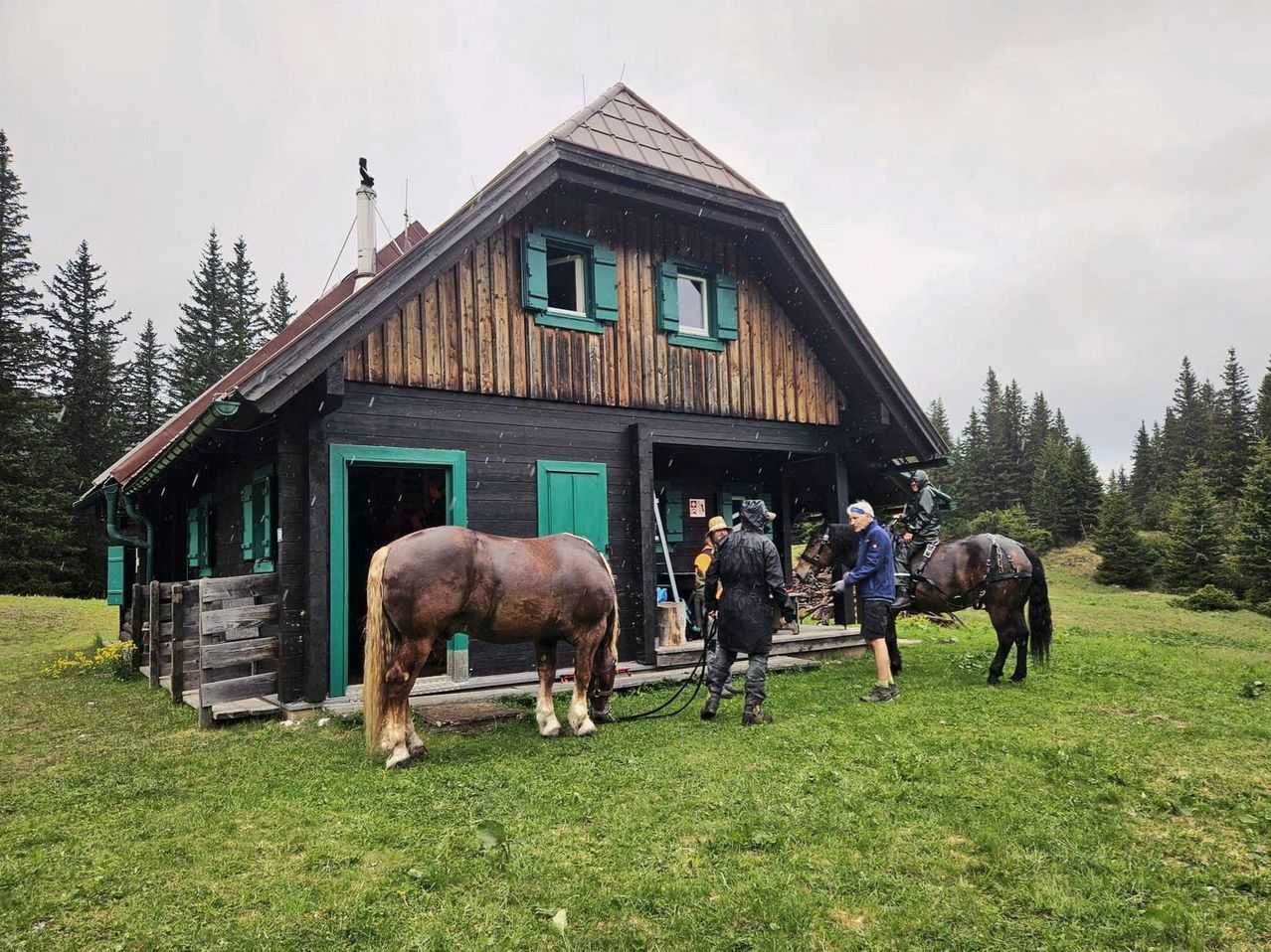 Three people are outside a wooden cabin with horses. One person is holding a rope attached to a horse. Another person is standing beside a horse. The third person is riding a horse.