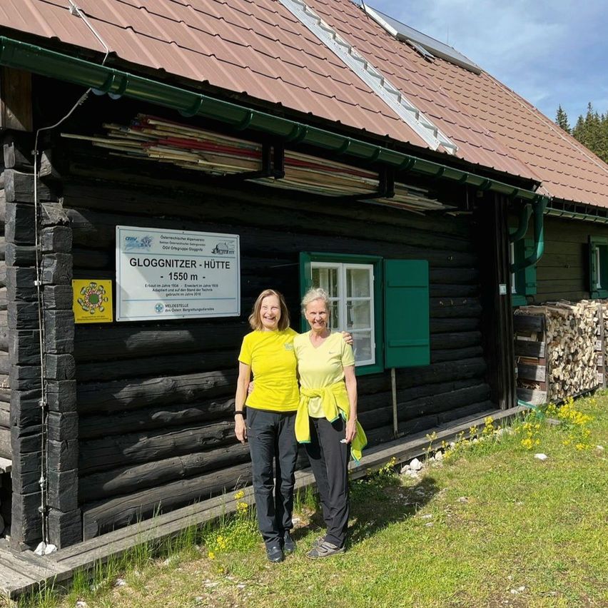 Two women stand in front of a log cabin with green shutters and a sign saying Gloggnitzer Hutte at 1550 meters. They are wearing yellow shirts and seem to be posing for a photo.