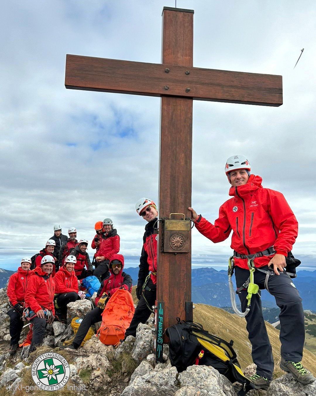 Eine Gruppe von Kletterern in roten Jacken und Helmen posiert am Gipfel mit einem hölzernen Kreuz.
