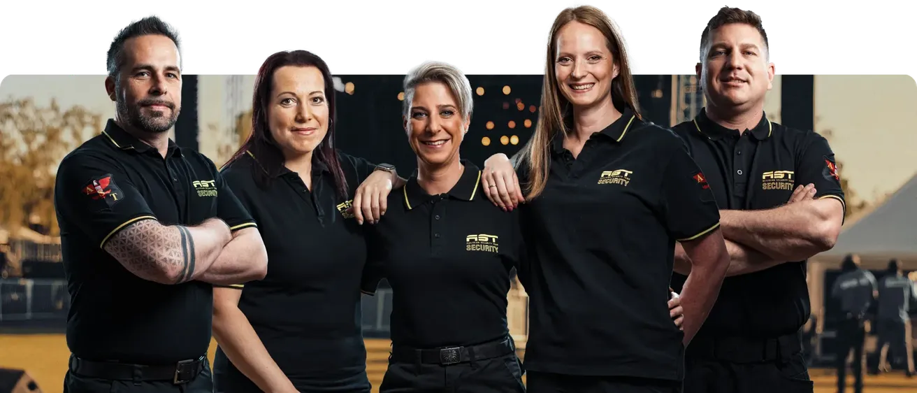 Drei Frauen in passenden schwarzen Sicherheitsuniformen lächeln für ein Foto. Sie tragen gelbe Akzente an ihren Kragen und Gürteln. Die Frau in der Mitte hat ihren Arm um die Schultern der anderen gelegt.