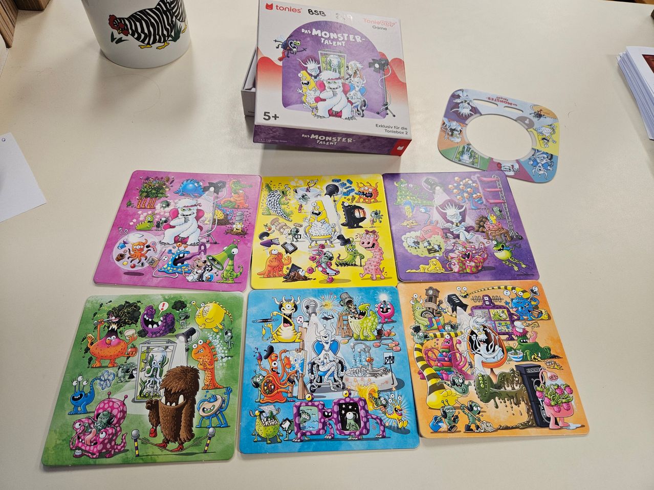 Auf einem Tisch ist eine Schachtel des 'Das Monster-Talente' Puzzlespiels für Kinder ab 5 Jahren zu sehen, mit farbenfrohen Illustrationen von Monstern. Neben der Schachtel befinden sich sechs lebhafte, monster-thematische Puzzleteile und ein rundes Puzzleteil daneben.