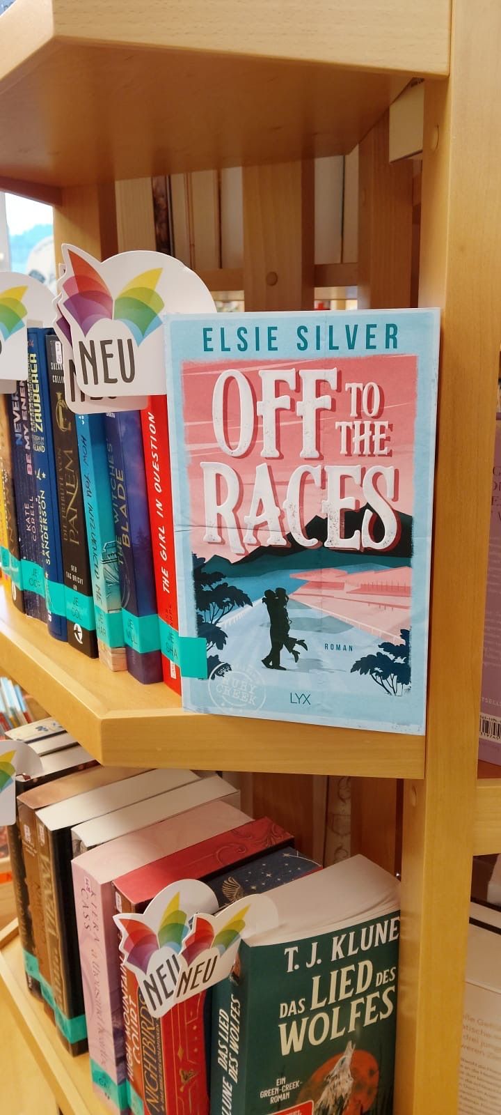Ein Buch mit dem Titel 'Off to the Races' von Elsie Silver auf einem Holzregal mit anderen Büchern und einem Schild, das 'NEU' anzeigt.
