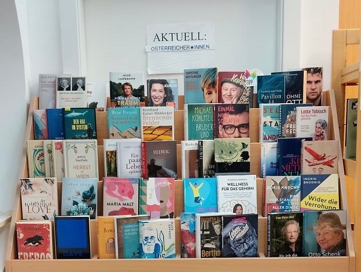 Ein hölzerne Bücherregal gefüllt mit verschiedenen Büchern. Ein weißes Schild mit schwarzen Buchstaben steht 'AKTUELL: OSTERREICHER*INNEN'. Die Bücher haben unterschiedliche Einbände, Titel und Autoren. Die Bücher sind in Reihen angeordnet.