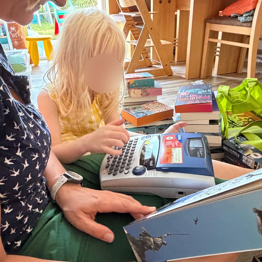 A young girl is using a typewriter while sitting on a woman's lap. They are surrounded by books and a bag.