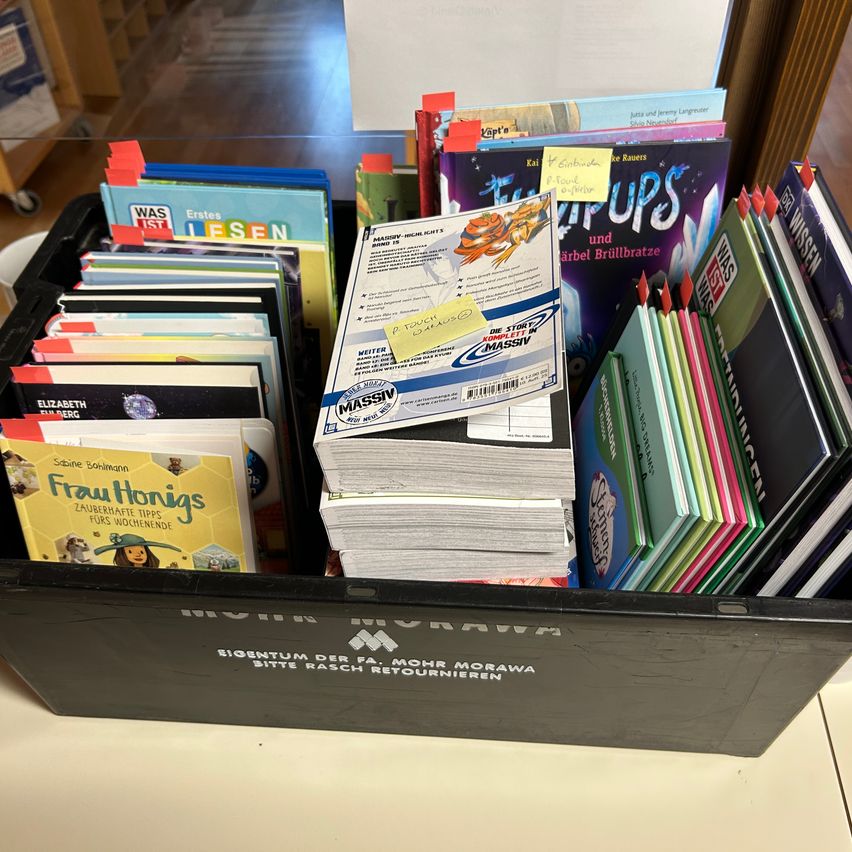 A black storage box filled with various books, some with sticky notes, arranged on a white surface. The books have different colors and titles, and some are stacked on top of each other. The box has a label that reads 'Eigntum der FA. Mohr Morawa Bitte rasch retournieren'.