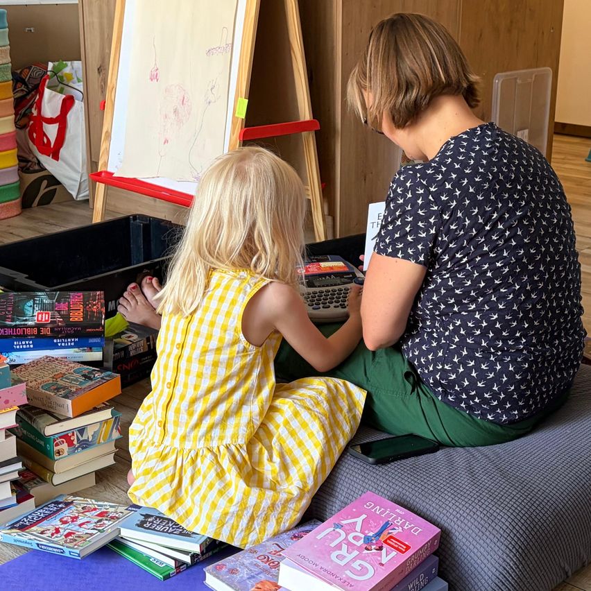 A woman and a young girl are sitting on the floor, surrounded by books. The woman is holding a calculator and appears to be teaching the girl. They are in a room with wooden floors and a wooden easel with a drawing on it.