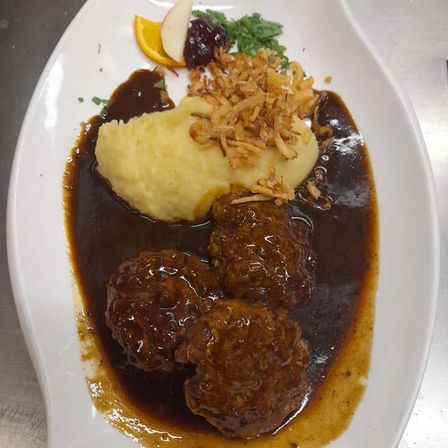 Ein Teller mit Fleischbällchen in brauner Soße mit Kartoffelpüree, Cranberry-Sauce und gebratenen Zwiebeln.