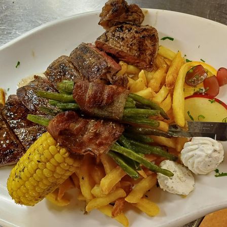 Ein weißer Teller mit Steak, Pommes, grünen Bohnen, Mais und Käse. Ein Messer liegt auf dem Teller.