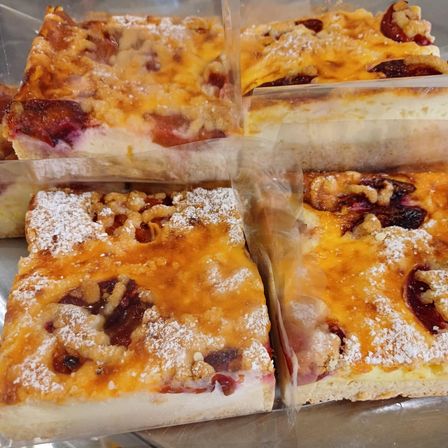 Mehrere Stücke von Obstkuchen mit einer goldbraunen Kruste, überzogen mit weißem Puderzucker und Walnussstücken, sind in durchsichtigen Plastikbehältern ausgestellt.