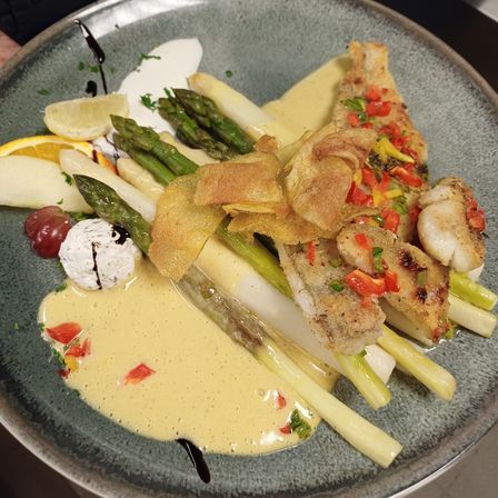 Ein Teller mit cremiger Sauce enthält Fischfilets, Spargel, eine Zitronenscheibe, eine Traube und Kartoffelchips. Es enthält auch rote Paprika und grüne Kräuter.
