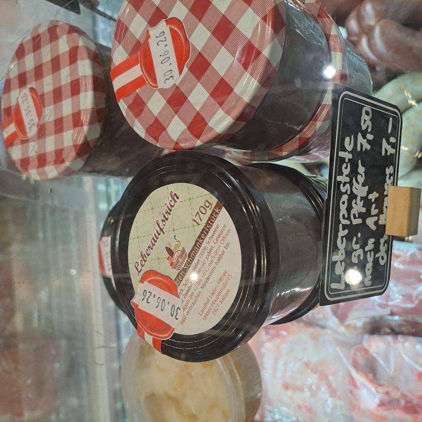 Drei Gläser mit rot-weiß karierten Deckeln stehen auf einer Glasvitrine. Ein schwarzes Glas mit weißem Etikett hat einen Preisschild mit '7,90' darauf. Andere Produkte sind im Hintergrund sichtbar.