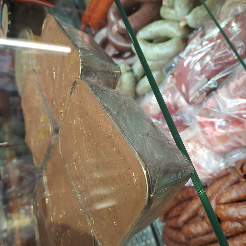 Drei Stücke Fleisch, in Folie eingewickelt, liegen in einer Glasvitrine. Im Hintergrund sind Würste und andere Fleischsorten zu sehen, alles ordentlich angeordnet.