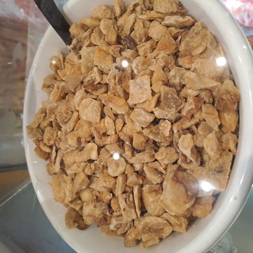 Eine weiße Schüssel gefüllt mit unregelmäßig geformten, bräunlich-gelblichen Stücken, die dehydriertes Obst oder Gemüse zu sein scheinen, mit einem schwarzen Löffel, der teilweise eingetaucht ist.