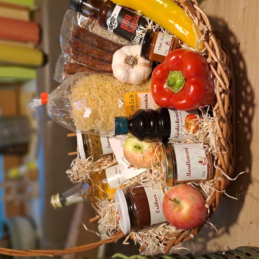 Ein Korb aus Weide enthält verschiedene Lebensmittel, darunter eine gelbe Paprika, eine rote Paprika, eine Flasche BBQ-Sauce, eine Knoblauchzehe, ein Glas Marmelade, eine Flasche Öl, ein Glas Konfitüre und mehrere Äpfel.