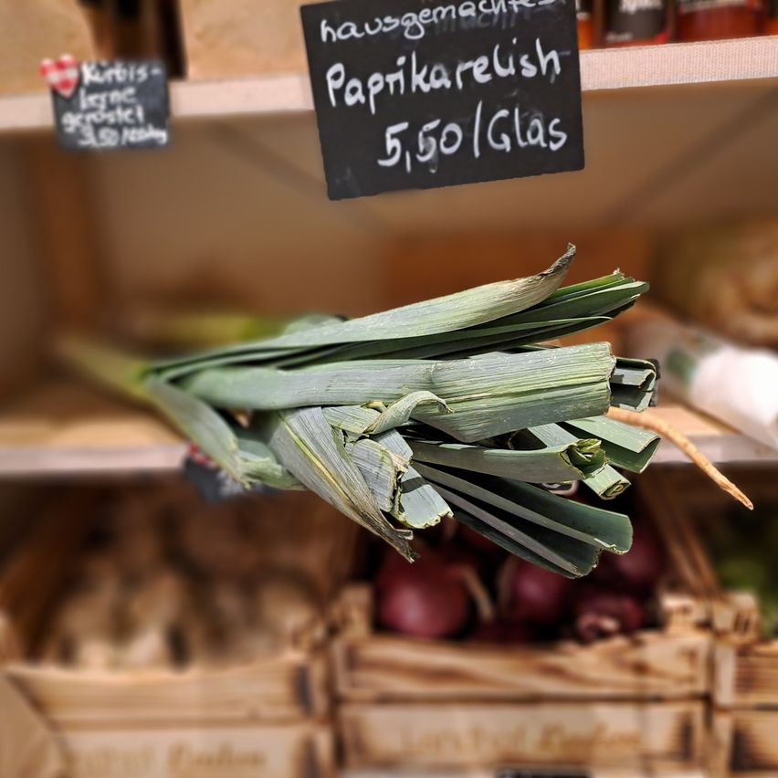 Ein Strauß grüner Zwiebeln hängt an einem Regal mit einem Preisschild. Dahinter sind Holzkisten und andere Gegenstände zu sehen.