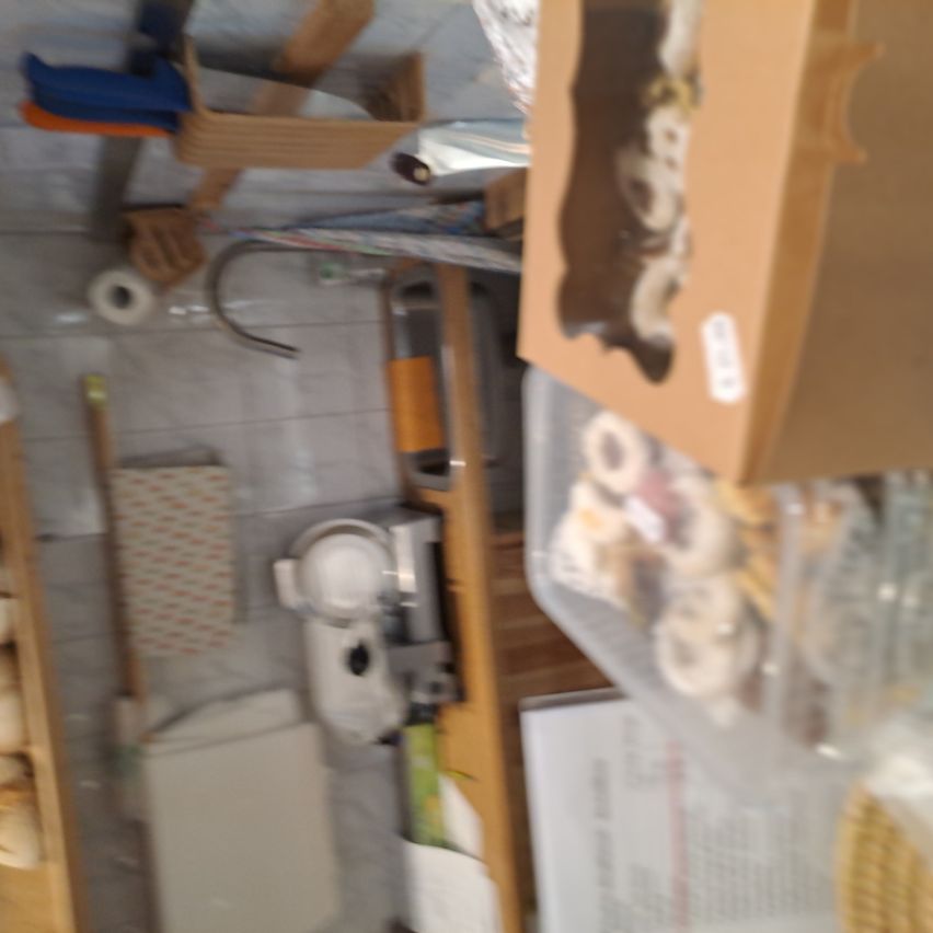 Ein überfüllter Raum mit einer Arbeitsplatte, einem Waschbecken und einem Holzschrank. Verschiedene Gegenstände wie eine Maschine, Aufbewahrungsboxen und eine Papierrolle sind verstreut.