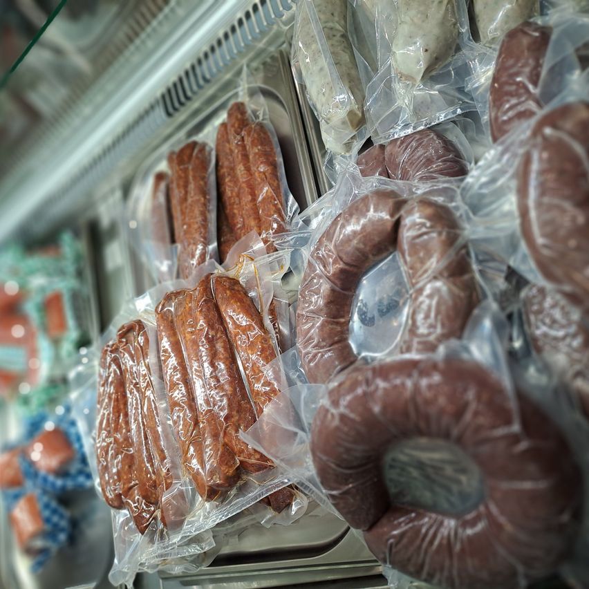 Verschiedene Würste in Plastikverpackungen auf einer gekühlten Anzeige. Mehrere Typen, einschließlich Hot Dogs und Würste, sind sichtbar. Die Anzeige ist in ordentlichen Reihen organisiert, und die Würste sind eng verpackt.