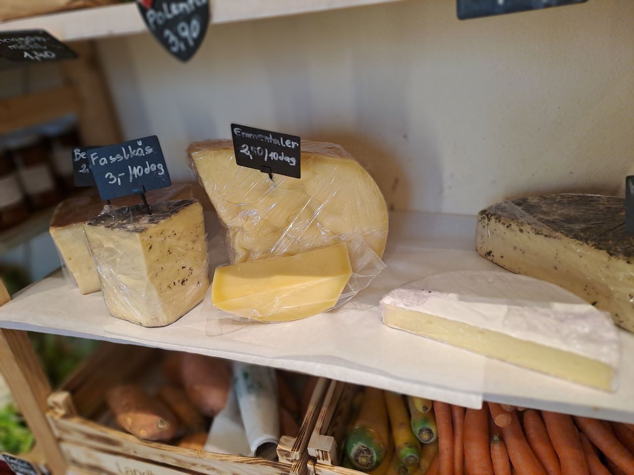 Eine Käseauswahl zeigt verschiedene Käsesorten, darunter ein Stück Emmentaler, ein Stück Fasslkas und andere. Sie sind auf einem Regal angeordnet, darunter Karotten und Süßkartoffeln.