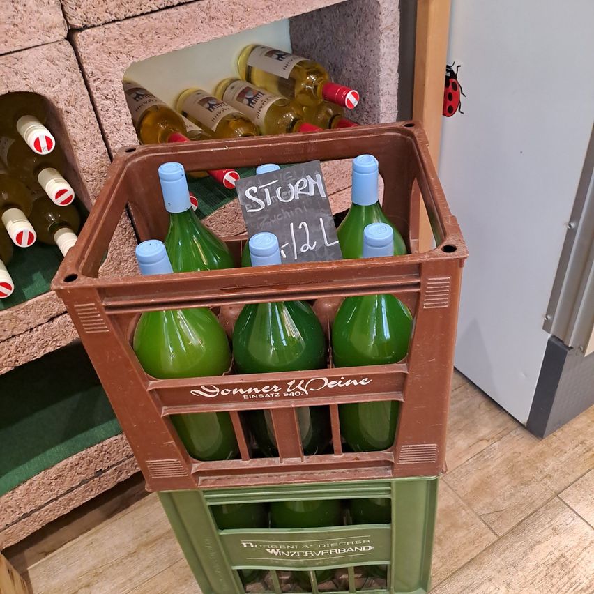 Ein brauner und grüner Kasten gefüllt mit grünen Weinflaschen, beschriftet mit 'STOR' und 'ZUCHI'. Der Kasten steht auf einem Holzboden mit einer Ziegelwand und einer Marienkäfer im Hintergrund.