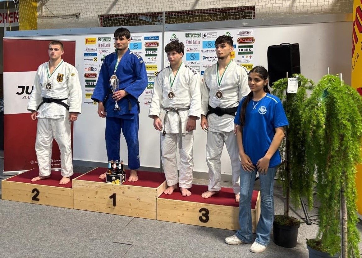 Fünf Männer stehen auf einem hölzernen Podium mit Medaillen um den Hals. Ein Mann hält einen Pokal und die anderen schauen auf etwas vor ihnen. Eine Frau in einem blauen T-Shirt steht neben ihnen.