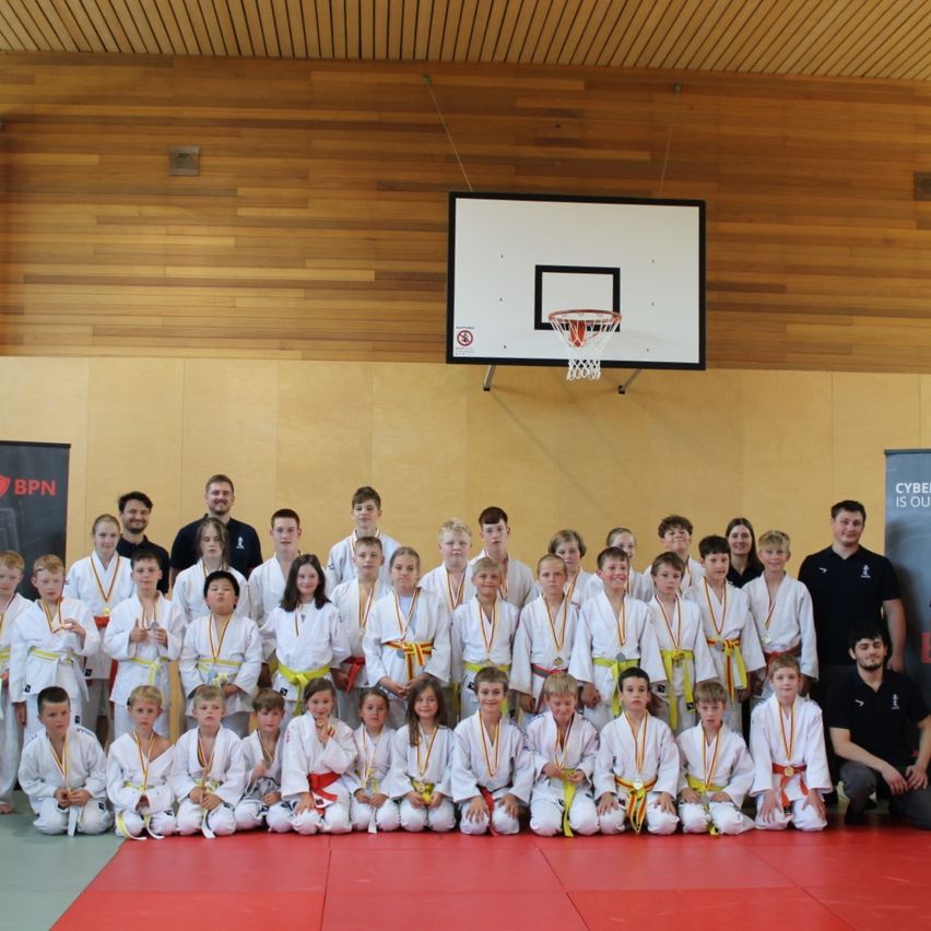 Eine Gruppe junger Kampfkünstler in weißen Uniformen mit Medaillen posiert für ein Foto in einer Turnhalle mit einem Basketballkorb an der Wand.