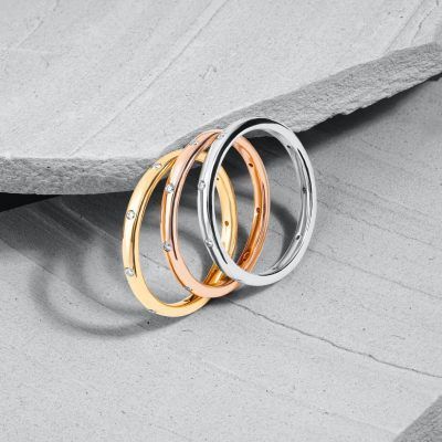 Drei metallische Ringe in verschiedenen Farben und mit kleinen Diamanten verziert sind auf einer grauen Oberfläche gestapelt.