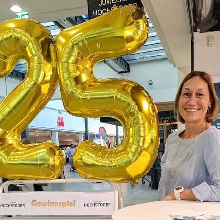 Eine lächelnde Frau steht neben einem großen goldenen Luftballon in Form einer '25', einem Tisch mit einem Schild, auf dem steht 'Danke, dass wir Teil Ihrer schönsten Momente sein dürfen'. Hinter ihr befindet sich ein Banner mit dem Text 'SOFO Sero'.