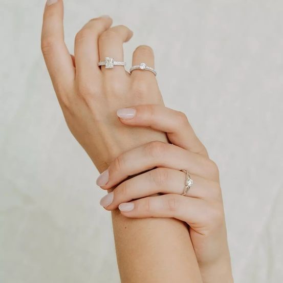 Eine Nahaufnahme von Händen mit drei Diamantringen an jedem Finger vor einem weißen Hintergrund.