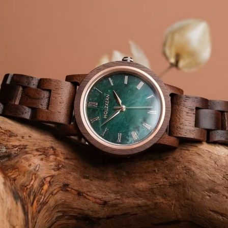 Eine Holzuhr mit grünem Zifferblatt und römischen Ziffern liegt auf einem Holzstück. Das Armband der Uhr ist braun.