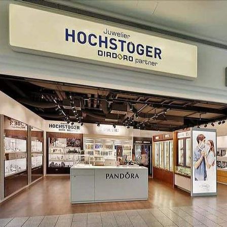 Der Eingang zu einem Juweliergeschäft mit einem großen Schild mit dem Wort 'Hochstöger'. Im Inneren befindet sich eine Theke mit dem Wort 'Pandora'. An der Wand befindet sich ein Poster mit einem Paar. Das Geschäft hat auch ein 'Swatch'-Schild.