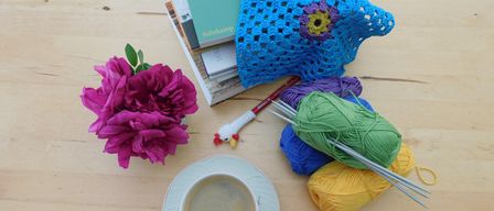 Eine Tasse Kaffee, Stricknadeln, Garn, ein gehäkeltes Stück und Bücher auf einem Holztisch. Eine rosa Blume ist links.