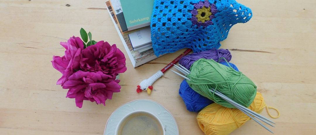 Eine Tasse Kaffee, Stricknadeln, Garn, ein gehäkeltes Stück und Bücher auf einem Holztisch. Eine rosa Blume ist links.