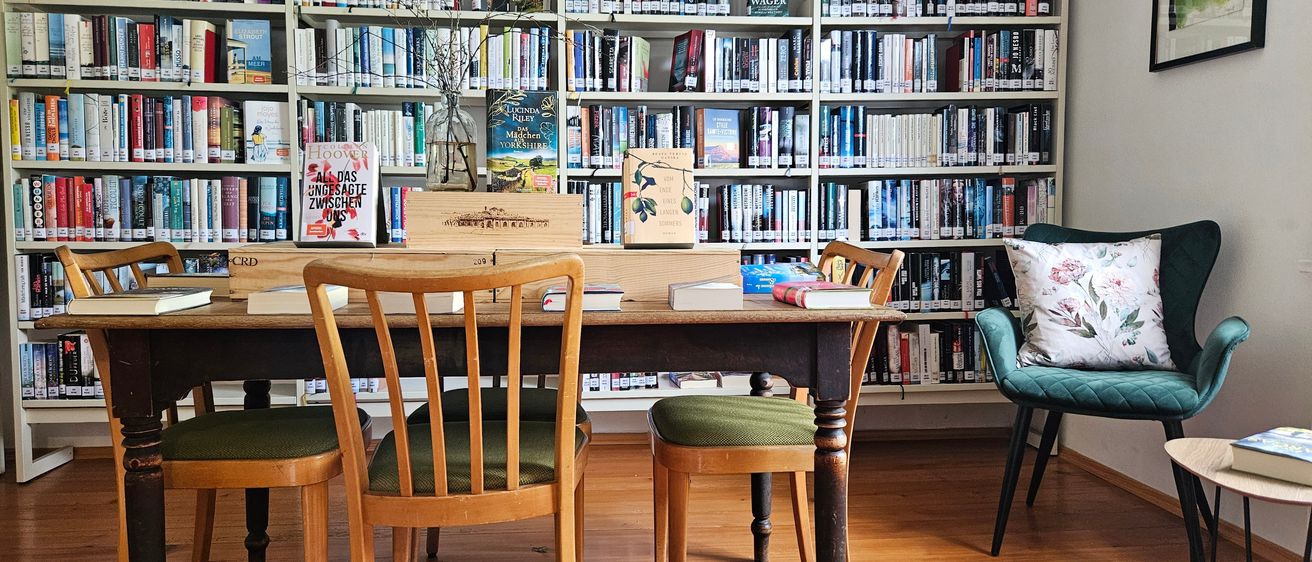 Ein gemütlicher Raum mit Holzboden, ausgestattet mit einem langen Holztisch und Stühlen, einem Regal voller Bücher und einem grünen Stuhl mit Kissen.