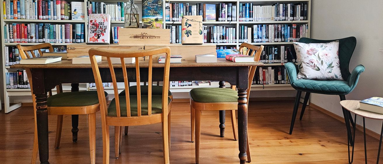 Ein gemütlicher Raum mit Holzboden, ausgestattet mit einem langen Holztisch und Stühlen, einem Regal voller Bücher und einem grünen Stuhl mit Kissen.