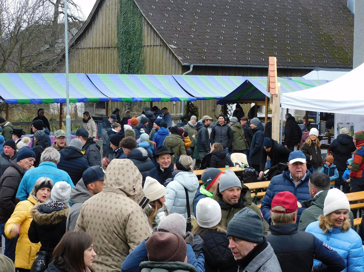 Eine Menschenmenge versammelt sich vor einem Holzgebäude, möglicherweise für eine Veranstaltung. Viele tragen Winterkleidung, einschließlich Mützen und Mänteln. Es gibt Tische und Zelte aufgestellt, was auf eine organisierte Versammlung hinweist.