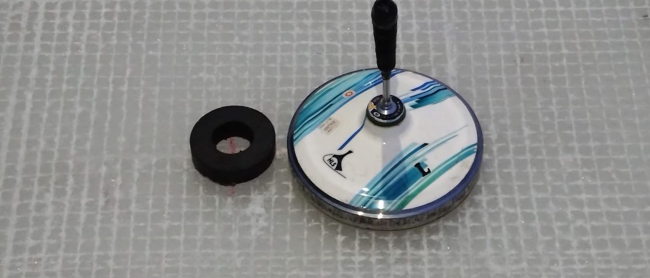 Ein rundes Objekt mit blau-weißem Design und schwarzem Griff neben einem schwarzen Gummiring auf einer gefliesten Oberfläche.