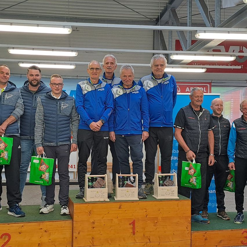 Eine Gruppe von Männern steht auf einer Bühne mit Trophäen. Sie tragen Sportbekleidung und einige halten Taschen. Sie lächeln und schauen in die Kamera. Hinter ihnen sind Banner.