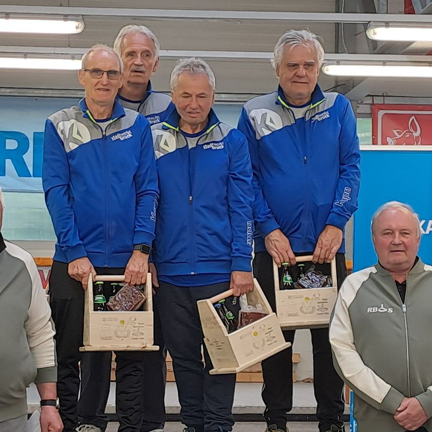 Fünf Männer stehen zusammen und halten Holzkisten mit Flaschen. Sie tragen passende blaue Jacken mit Logos und scheinen für ein Foto zu posieren. Der Hintergrund zeigt ein Banner mit einem großen Buchstaben 'R'.