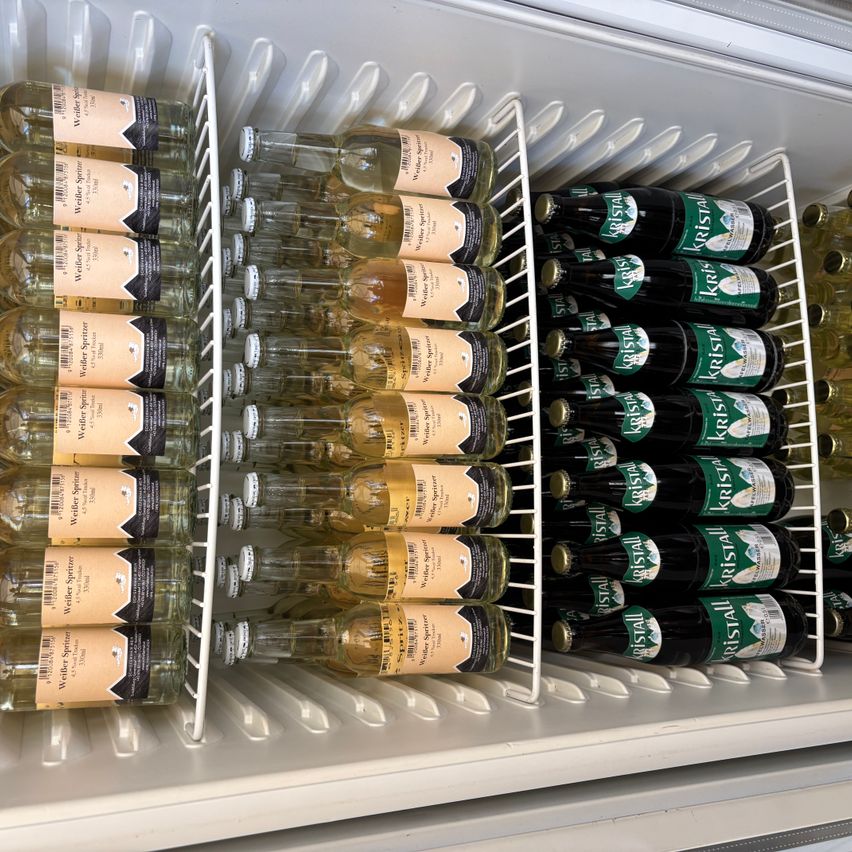 Ein offener Kühlschrank mit weißen Regalen enthält mehrere Alkoholflaschen. Links befinden sich Flaschen von 'Weinberger Spätlese' und rechts Flaschen von 'Kristall'. Die Flaschen sind in Reihen mit unterschiedlichen Mengen angeordnet.