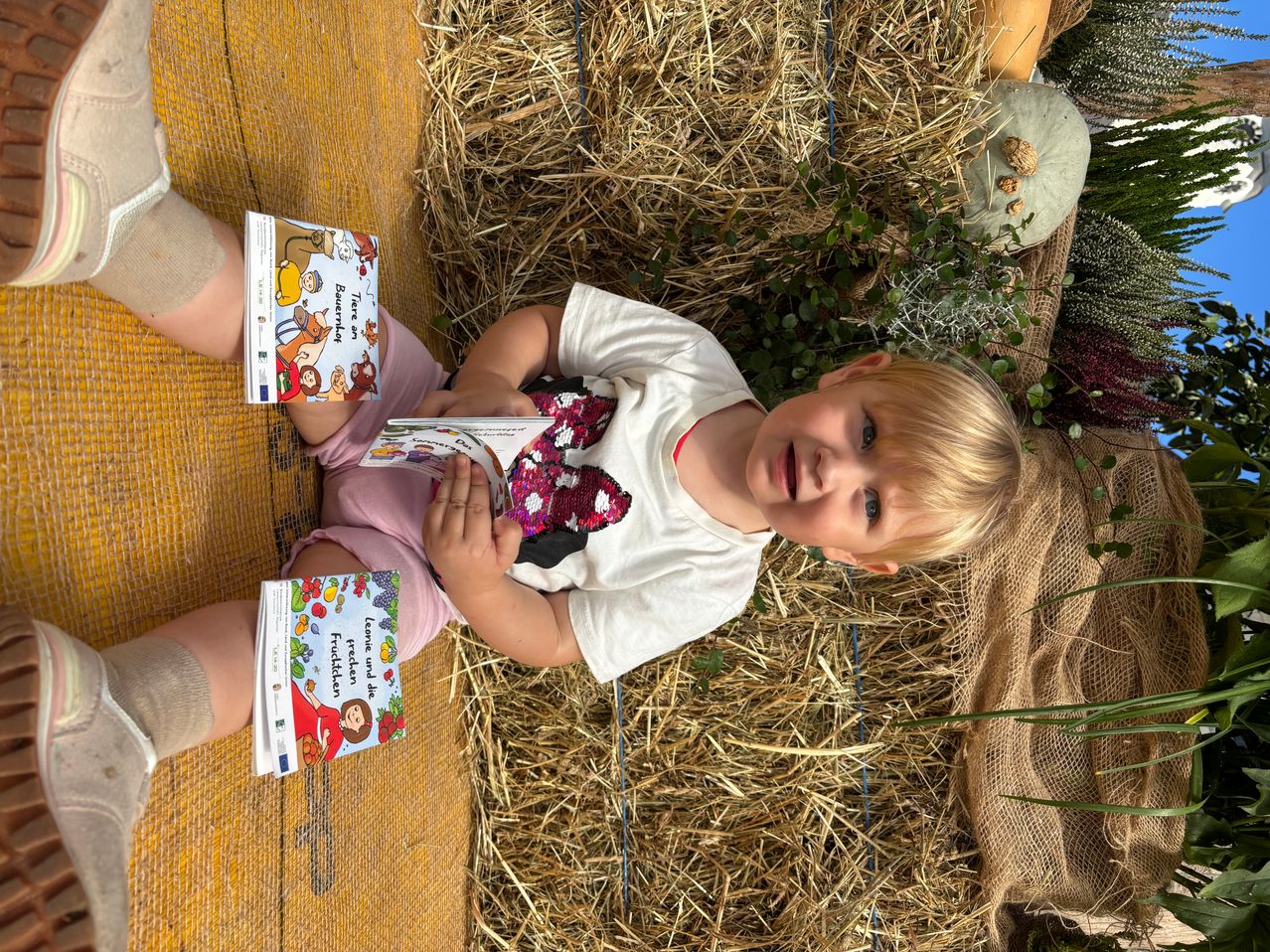 Ein Kind liegt lächelnd auf einem Heuballen und schaut in die Kamera. Das Kind trägt ein rosa Kleid und hält ein Buch mit einer Comicfigur.
