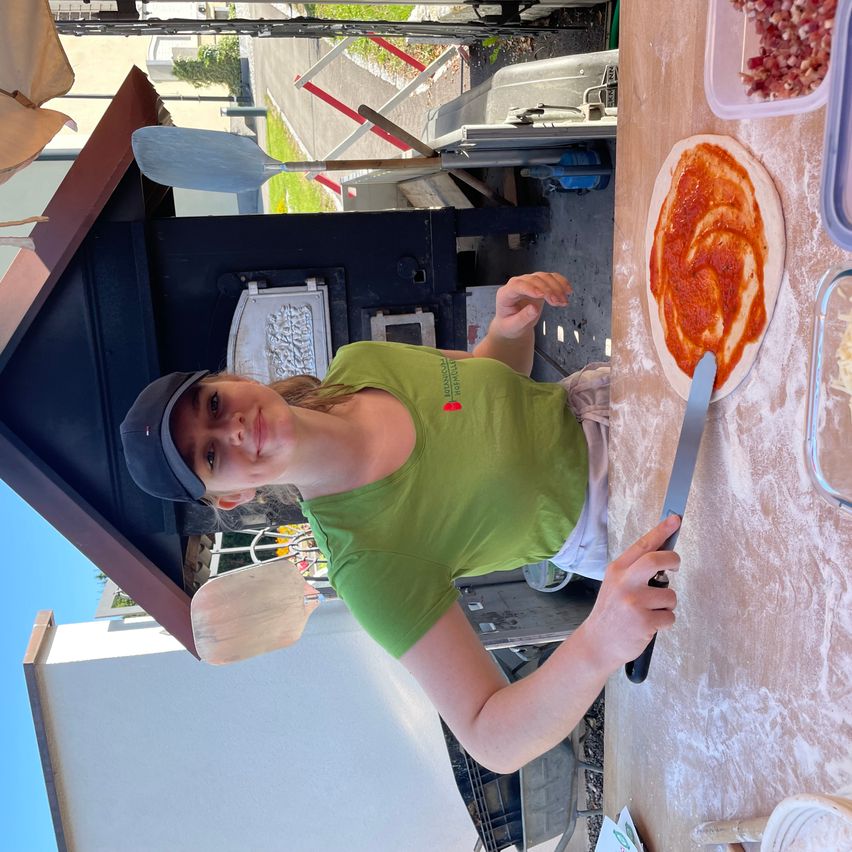 Eine Frau in einem grünen Shirt und einer Kappe bereitet eine Pizza mit Tomatensoße, Käse und Fleisch auf einem Holztisch zu. Im Hintergrund sind ein Pizzaofen und ein Gebäude zu sehen.