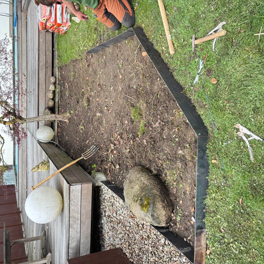 Ein Gärtner kniet auf dem Rasen, umgeben von Werkzeugen, Steinen und einem Holzzaun. Ein Stein liegt in der Mitte des vorbereiteten Bodens.