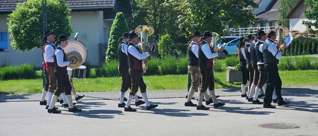 Eine Gruppe von Männern in traditioneller deutscher Kleidung marschiert in einer Parade und spielt Blechblasinstrumente vor einem Haus mit braunem Dach.